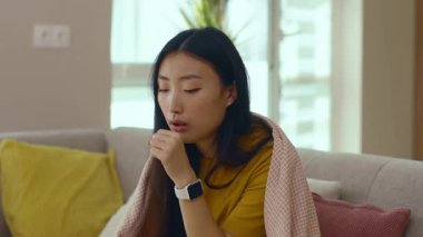 Kanepede oturan, battaniyeli, öksürük hastası Asyalı genç kadın kendini evinde hissediyor. Salgın. Coronavirus. Hastalıklı hastalık stresle karşı karşıya. Hastalık. Yavaş çekim