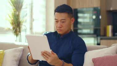 Ciddi Asyalı genç adam evdeki kanepede oturan tablet bilgisayar kullanıyor. Çinli iş adamı çok yakışıklı. İletişim. Kapatın. Yavaş çekim