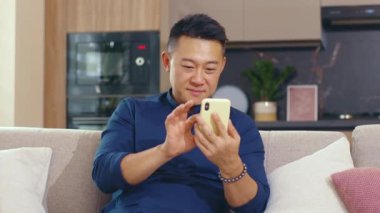 Gülümseyen mutlu Asyalı genç adam evdeki koltukta otururken telefon kullanıyor. Akıllı telefon bağlantısı iş adamı sosyal medyada sohbet ediyor. Kapatın. Portre. Yavaş çekim