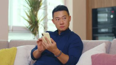 Asyalı ciddi bir genç adam telefon kullanıyor. Kanepede oturup evi kolaçan ediyor. Akıllı telefon bağlantısı iş adamı sosyal medyada sohbet ediyor. Kapatın. Portre. Yavaş çekim