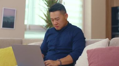 Kanepede oturan mutsuz Asyalı adam evde dizüstü bilgisayar kullanıyor. Ağ işi üzgün adam iç teknoloji sosyal medya. Yavaş çekim