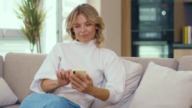 Mutlu sarışın kadın telefon sosyal medya platformunu kullanıyor ve evdeki kanepede oturuyor. İnternet kanepesi. Evin içinde güzel bir mesaj. Kapatın. Yavaş çekim