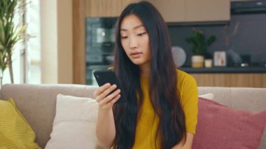 Çekici Asyalı genç kadın evdeki koltukta otururken telefon kullanıyor. Sosyal medya mesajları uzak iletişimin tadını çıkarıyor. Sosyal medya. Kapatın. Yavaş çekim