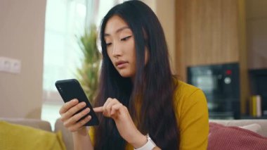 Çekici Asyalı genç kadın telefonuyla evdeki kanepeye oturuyor. Sosyal medya mesajları uzak iletişimin tadını çıkarıyor. Sosyal medya. Kapatın. Yavaş çekim