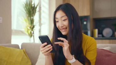Gülümseyen Asyalı genç kadın evdeki koltukta oturan telefonu kullanıyor. Sosyal medya mesajları uzak iletişimin tadını çıkarıyor. Sosyal medya. Kapatın. Yavaş çekim