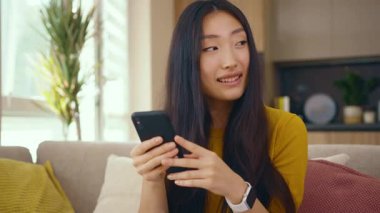 Mutlu Asyalı genç kadın evdeki koltukta otururken telefonu kullanıyor. Sosyal medya mesajları uzak iletişimin tadını çıkarıyor. Sosyal medya. Kapatın. Yavaş çekim
