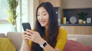 Gülümseyen Asyalı genç kadın telefonuyla evdeki kanepeye oturuyor. Sosyal medya mesajları uzak iletişimin tadını çıkarıyor. Sosyal medya. Yavaş çekim