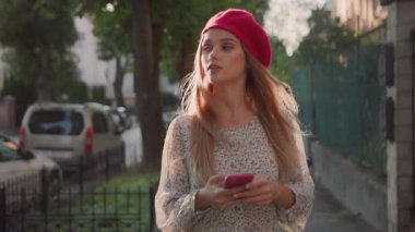 Neşeli Kafkasyalı güzel sarışın bayan şık giyinmiş kırmızı bere elinde telefonla güneşli bir şehirde arama yapıyor..