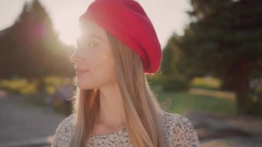 Genç, güzel, sarışın, güzel kırmızı bereli bir kadın kameraya gülümsüyor. Günbatımı arka planı. Güneşli güzel bir gün. İnsanlar.