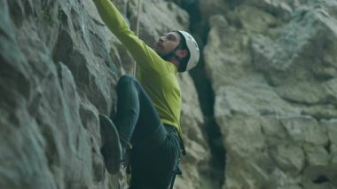 Kafkasyalı genç hippi dikkatlice kayaya tırmanıyor, sıkıca kavrıyor ve yukarı bakıyor. Sakallı sporcu. Aşırı hareketlilik. Yürüyüş.