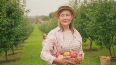 Yaşlı kadının portresi kırmızı elmalarla dolu sepeti tutuyor. Arka planda büyükbabası ve güzel torunları bahçede yürürken. Çocukluk, yemek, köy. Gün ışığında. Yavaş çekim