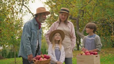 Sonbahar bahçesinde neşeli bir aile görüntüsü. Gülümseyen çiftçiler güneşli bahçede kırmızı elma topluyor. Büyükbaba şapkalı güzel bayanı dikkatle dinliyor, birlikte vakit geçiriyorlar. Açık havada. Organik meyveler.