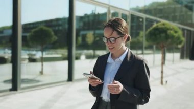 Zarif, kendine güvenen, zarif, resmi takım elbiseli beyaz bir kadının akıllı telefonu elinde tutarak etrafa bakındığı görüntüler. Bun saç stiliyle gülümseyen zeki iş kadınının portresi. Arka plan bulanık. Açık havada.