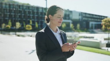 Kendine güvenen, güzel görünümlü genç bir kadının akıllı telefonu elinde tutarak sohbet ettiği görüntüler. Kameraya bakan çekici iş kadınının portresi. Modern binaların bulanık arkaplanı