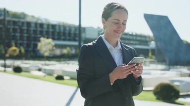 Genç, arkadaş canlısı, beyaz bir kadını gülerken, mesaj okurken, öğle arasında akıllı telefondan mesajlaşırken çekmek. Modern binaların bulanık arkaplanı. Açık havada. Çalış. Gündüz vakti. Sonbahar. Şehre. İletişim