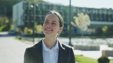 Etrafa bakıp akıllı telefon kullanan, arkadaşlarıyla mesajlaşan, gülümseyen beyaz iş kadınının yakın çekim görüntüleri. Resmi giysiler içinde kendine güvenen hoş görünümlü bir kızın portresi. Açık havada. Ofis binasının dış tasarımı