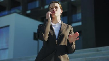 İş kıyafetli, yakışıklı, ciddi bir kadının yatay görüntüsü. Takım elbiseli, kendine güvenen meşgul bir kızın ofis merkezinde akıllı telefondan konuştuğu görüntüler. Açık havada. Günlük hayat. Her zamanki gibi. Şehir tasarımı