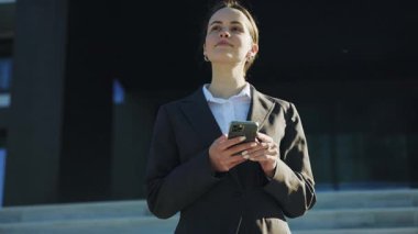 Dostane görünüşlü, gülümseyen, siyah takım elbiseli, beyaz bir kadın. Şık saç stili ile dalgın ve çekici bir kız ofis binasının yakınında akıllı telefon kullanıyor. Açık havada. Şehre. Yaz zamanı. İletişim
