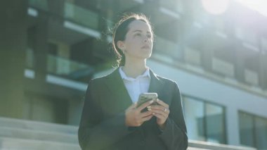 Düşünceli, yakışıklı, beyaz bir kadının portresi gözlerini kaçırıyor, akıllı telefondan mesajlaşıyor. İş gezisi sırasında modern otelin yakınındaki çekici kızın yatay görüntüsü. Açık havada. Güneş ışığı. Sonbahar