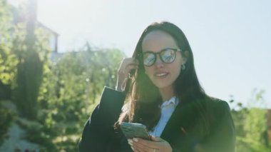 Elinde cep telefonu olan, sohbet eden, yakışıklı, beyaz bir kadının fotoğrafı. Akıllı telefon ekranına bakan kendine güvenen muhteşem iş kadınının portresi. Güneşli bir gün. Açık havada. Sonbahar. Güneş ışığı
