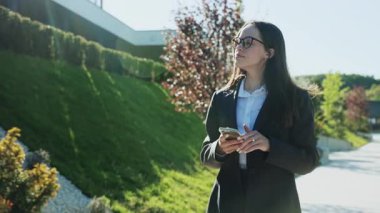 Parkta yürüyen, öğle arasında etrafa bakınan güzel beyaz bir kadının portresi. Akıllı telefon kullanan güzel bir kızın görüntüsü. Gündüz vakti. Çalışma günü