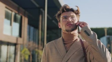 Gözlüklü yakışıklı adam telefonla konuşuyor. Etrafına bak ve caddede dur. Gün ışığında ciddi bir çocuk. Portre. Yavaş çekim