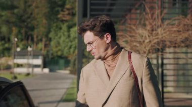 Kapıları açıp sürücü koltuğunda oturan gözlüklü çekici bir adam. Modern sokak. Güneş ışığı. Sıradan bir tarz. Yavaş çekim