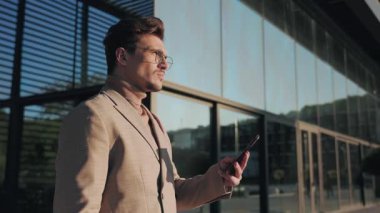 Şık bir adam, güneşli bir günde şık ve modern bir cam binanın önünde dikilirken akıllı telefonuna özgüvenle bakar.
