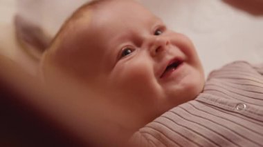 Bej takım elbiseli şirin bir bebeğin yakın plan çekimleri ahşap bir karyolada uzanmış, ebeveynlere bakıyor, gülümsüyor ve gülüyor. Mutluluk. Çocuk odasında yeni doğmuş sevimli bir bebeğin portresi. İçeride, evde.