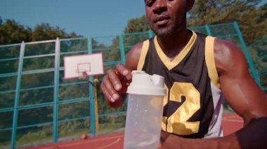 Afro-Amerikan sokak oyuncusu açık hava oyun parkında su molası veriyor. Güneş ışığı var. Yaz. Spor. Antrenman. Mahkeme. Yavaş çekim