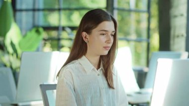 Çekici, çalışan, bilgisayarını kullanarak kameraya bakan, gülümseyen genç bir kadın aydınlık ofiste mutlu hisseder. Modern çalışma alanı. Teknoloji. Girişimci. İş kadınıyım. Yavaş çekim