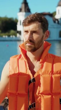 Parlak turuncu bir can yeleği giyen bir adam gururla, büyüleyici bir su manzarasının yanında duruyor.