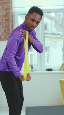 Bir adam, iyi aydınlatılmış bir odada parlak sarı bir direniş bandı kullanarak detaylı bir egzersiz rutini gösteriyor.