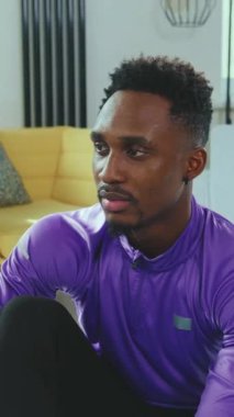 Canlı mor bir atletik gömlek giymiş fit bir adam modern bir oturma odasında düşünceli ve düşünceli bir şekilde oturur.