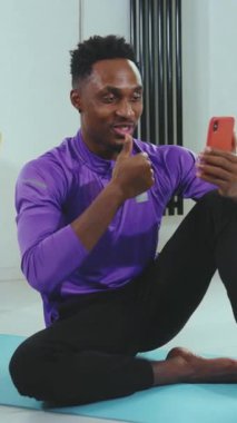 Spor kıyafeti giymiş bir adam yoga minderinde spor yaparken akıllı telefonuyla etkileşime girerken görülüyor.