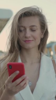 Uzun dalgalı saçları olan genç bir kadın deniz kenarında eğlenirken akıllı telefonunu kullanırken neşe saçıyor.