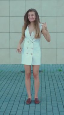 Şık, beyaz bir elbise giymiş neşeli ve neşeli genç bir kadın modern bir arkaplanda neşe içinde poz veriyor.