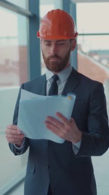 Sıkı bir şapka takmış iyi giyimli bir kişi modern çalışma alanındaki inşaat belgelerini özenle inceliyor.