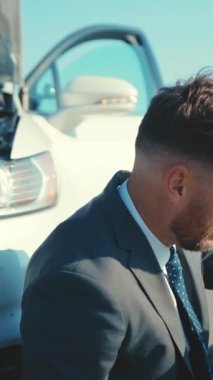 Terzi kıyafeti giymiş bir işadamı, açık mavi gökyüzünün altında, parlak bir aracın yanında güvenle duruyor.