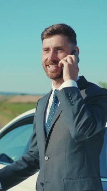 Şık takım elbiseli kendine güvenen bir adam telefonuyla güneşli bir ortamda bir arabanın yanında konuşuyor.