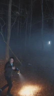 Gölgeli bir figür sisli bir ormanda dikkatlice geziniyor, etraftaki garip ve ürkütücü ışıklarla aydınlatılıyor.