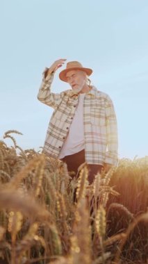 Bir adam emin adımlarla ayakta duruyor uçsuz bucaksız altın bir buğday tarlasında hasır şapka takmış ve gün batımında rahat bir yaz elbisesi giymiş.