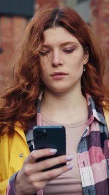 Kıvırcık kızıl saçlı genç bir kadın akıllı telefonuna odaklanmış, şehir çevresiyle iç içe.