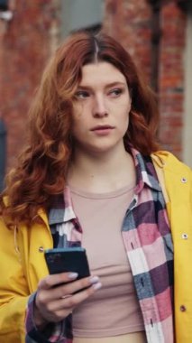 Dalgalı kızıl saçlı genç bir kadın açık havada eğlenirken akıllı telefonuyla meşgul oluyor.