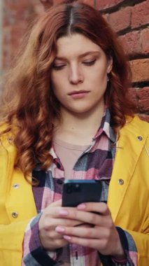 Parlak sarı ceket giyen genç bir kadın şehir ortamında telefonuna odaklanmış durumda.