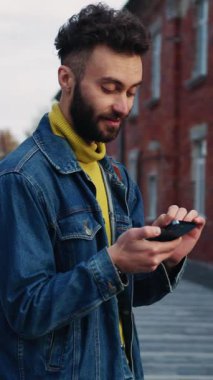 Genç bir adam, şehir merkezindeki tuğla binaların önünde dururken akıllı telefonuyla meşgul.