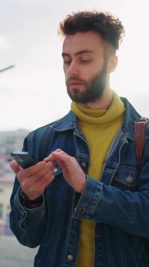 Şık bir genç adam gün ışığında duruyor, akıllı telefonunu kontrol ediyor, modern teknoloji ve moda trendlerini vurguluyor.