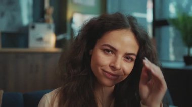 Kameraya gülümseyen kahverengi gözlü çekici genç bir kadının yakın plan portresi. Güzel model uzun kıvırcık saçlara dokunuyor. Bulanık arka planda oturma odası