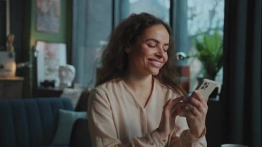 Akıllı telefon kullanan, başka tarafa bakan çekici, hayalperest, beyaz bir kadının portresi. Uzun kıvırcık saçlı, elinde cep telefonuyla evde sohbet eden güzel bir kız.