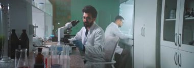 Gözlüklü ve koruyucu eldivenli gülümseyen bir bilim adamı mikroskop altında laboratuvarda bir tüp tutuyor. Tıp, biyoteknoloji, kimya. Arka planda iş arkadaşım yürüyor.